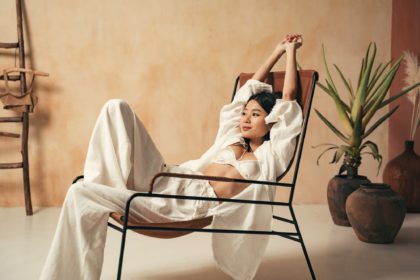 Asian female in rustic minimalistic decor sitting on lounger with raised hands stretching after nap