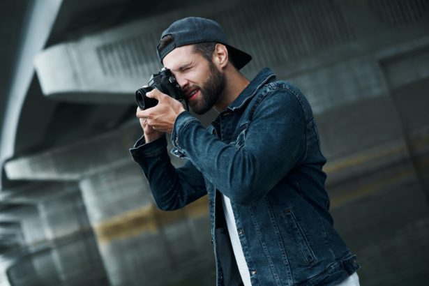 Photography hobby. Young stylish man standing on city street taking photos on camera concentrated