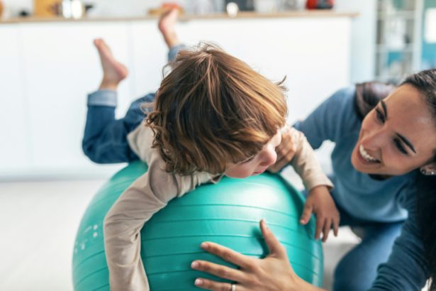 Young beautiful mother playing with fitness ball on the floor with her son in living room at home.